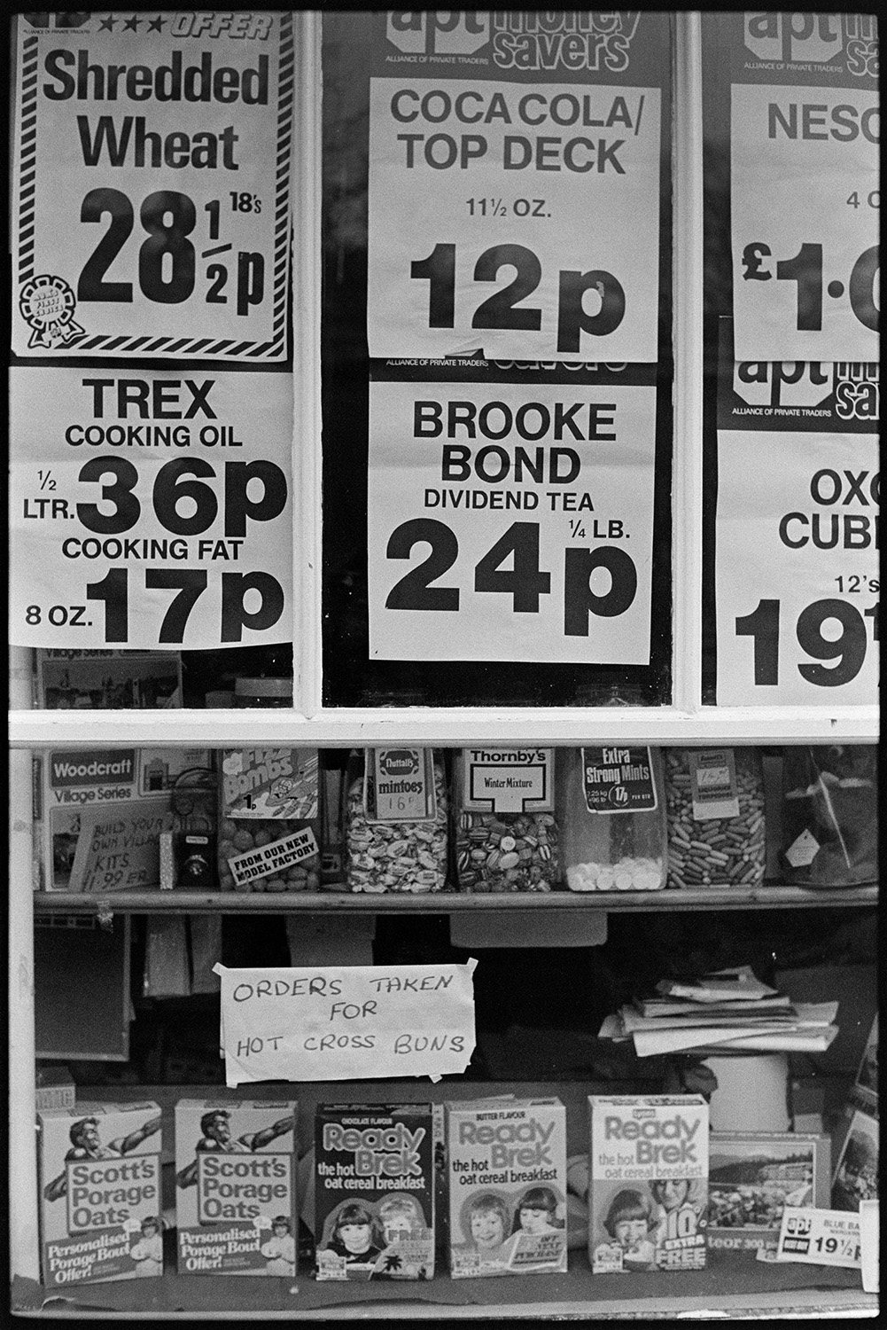 Grocery shop window with price signs and notice, hot cross buns. 
[The shop front window of London House Stores in Fore Street, Dolton. Posters advertising shredded wheat, coca cola and cooking oil, amongst other goods, are stuck in the window. Sweet jars, porridge boxes and a sign reading 'Orders Taken for Hot Cross Buns' are also visible.]