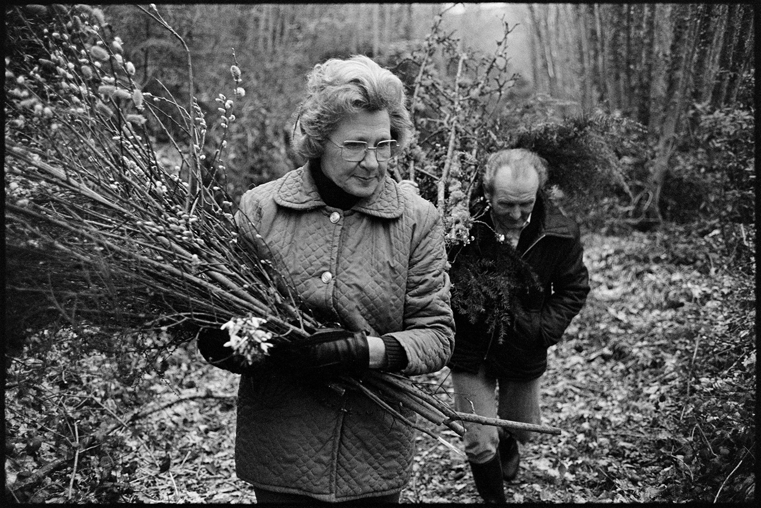 Couple collecting branches and Pussy Willow for Easter decorations in church. 
[Beatrice Nancekivell and Tom Nancekivell carrying branches of pussy willow along a path by a wooded area at Combehouse, Dolton, to use in Easter decorations at the Church.]