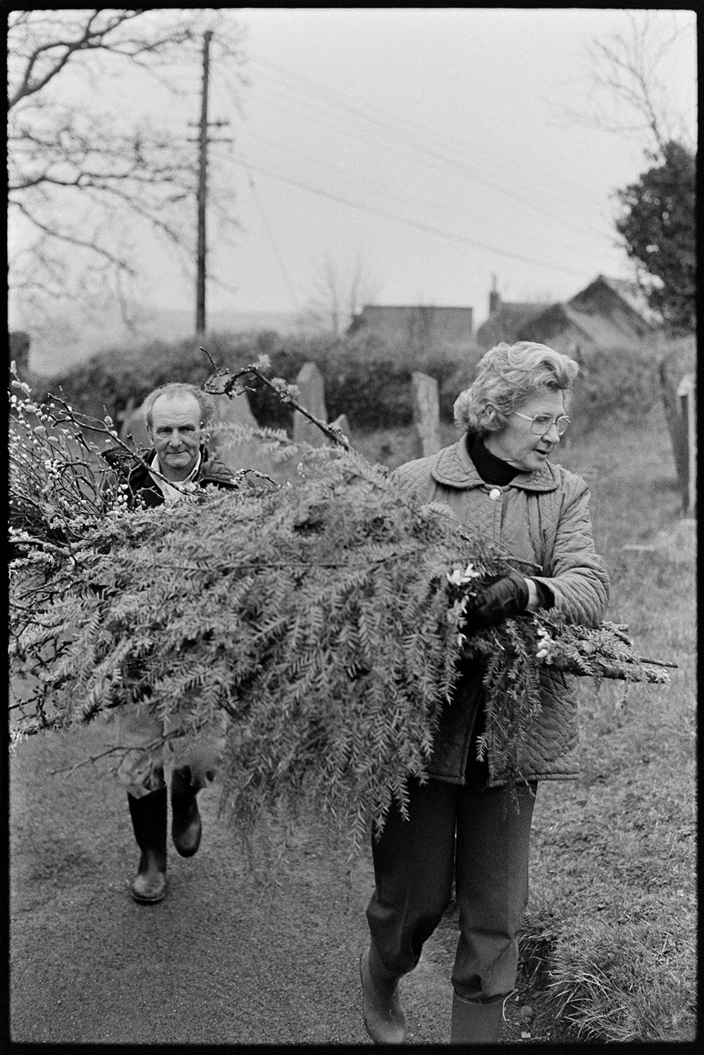 Couple collecting branches and Pussy Willow for Easter decorations in church. 
[Beatrice Nancekivell and Tom Nancekivell carrying branches of pussy willow and trees along the church path at Dolton Church, to use for Easter decorations in the Church. Gravestones can be seen in the background.]