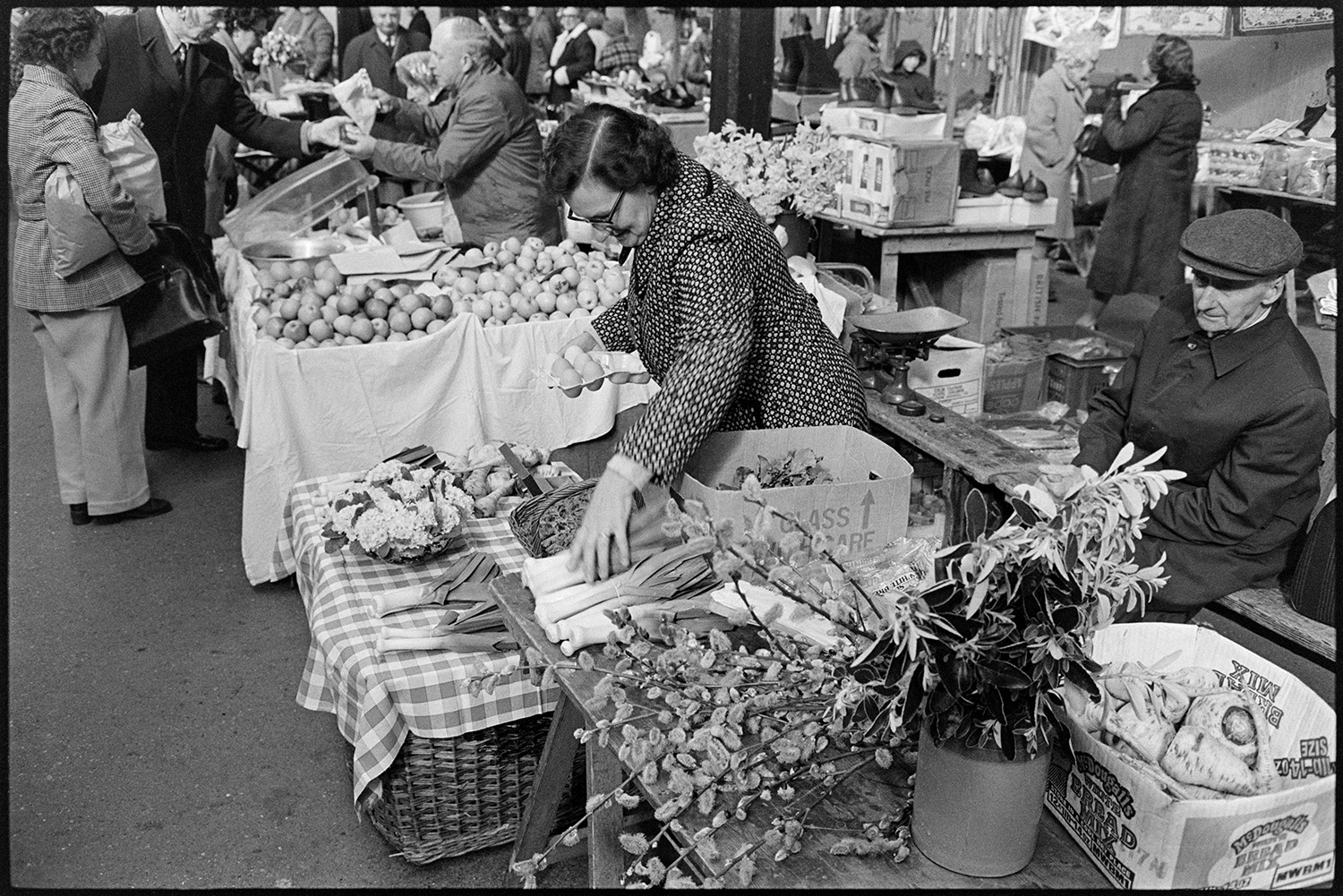 Stalls at Pannier market. Eggs, vegetables, flowers.
[A woman and man running a vegetable stall at Barnstaple Pannier Market. They are selling leeks, flowers and parsnips, amongst other goods. Customers and other stalls can be seen in the background.]