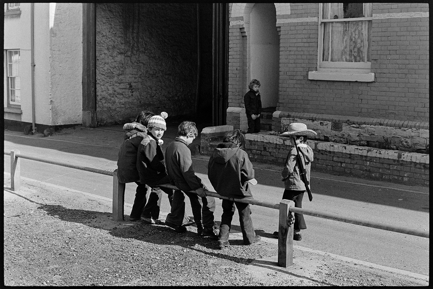 Street scenes. Children and old houses before development.
[Children sitting on a rail by Tuly Street, Barnstaple. One boy is waiting at a door on the opposite side of the street, and another boy is wearing a hat and carrying a toy gun.]