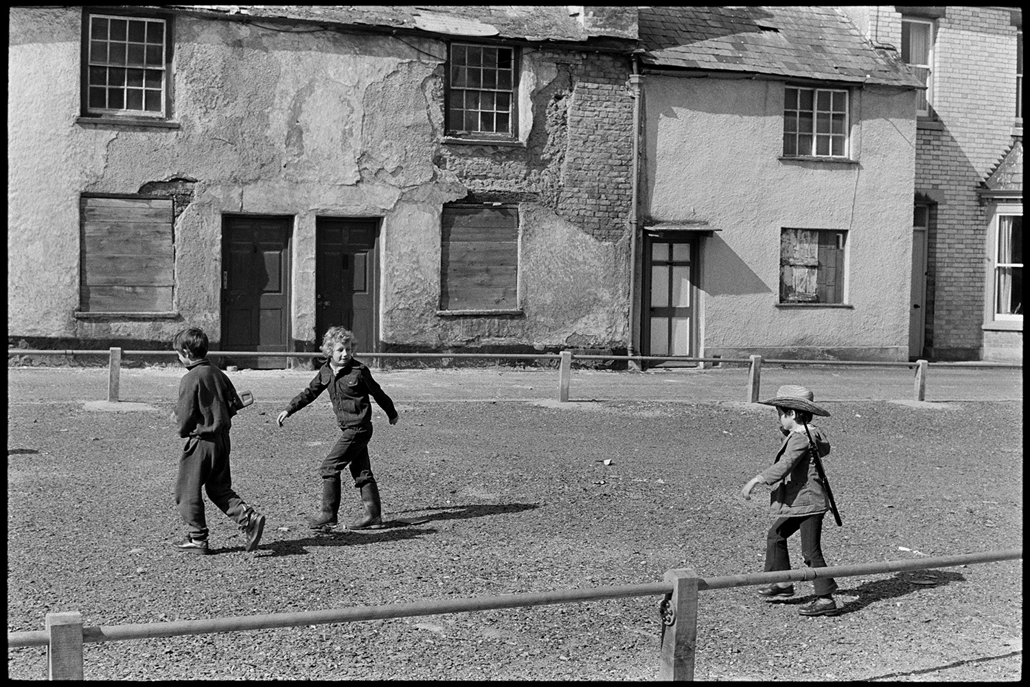 Street scenes. Children and old houses before development.
[Three children, one wearing a hat and carrying a toy gun, playing in the street at Tuly Street, Barnstaple. Houses with boarded up windows are visible on the other side of the street.]