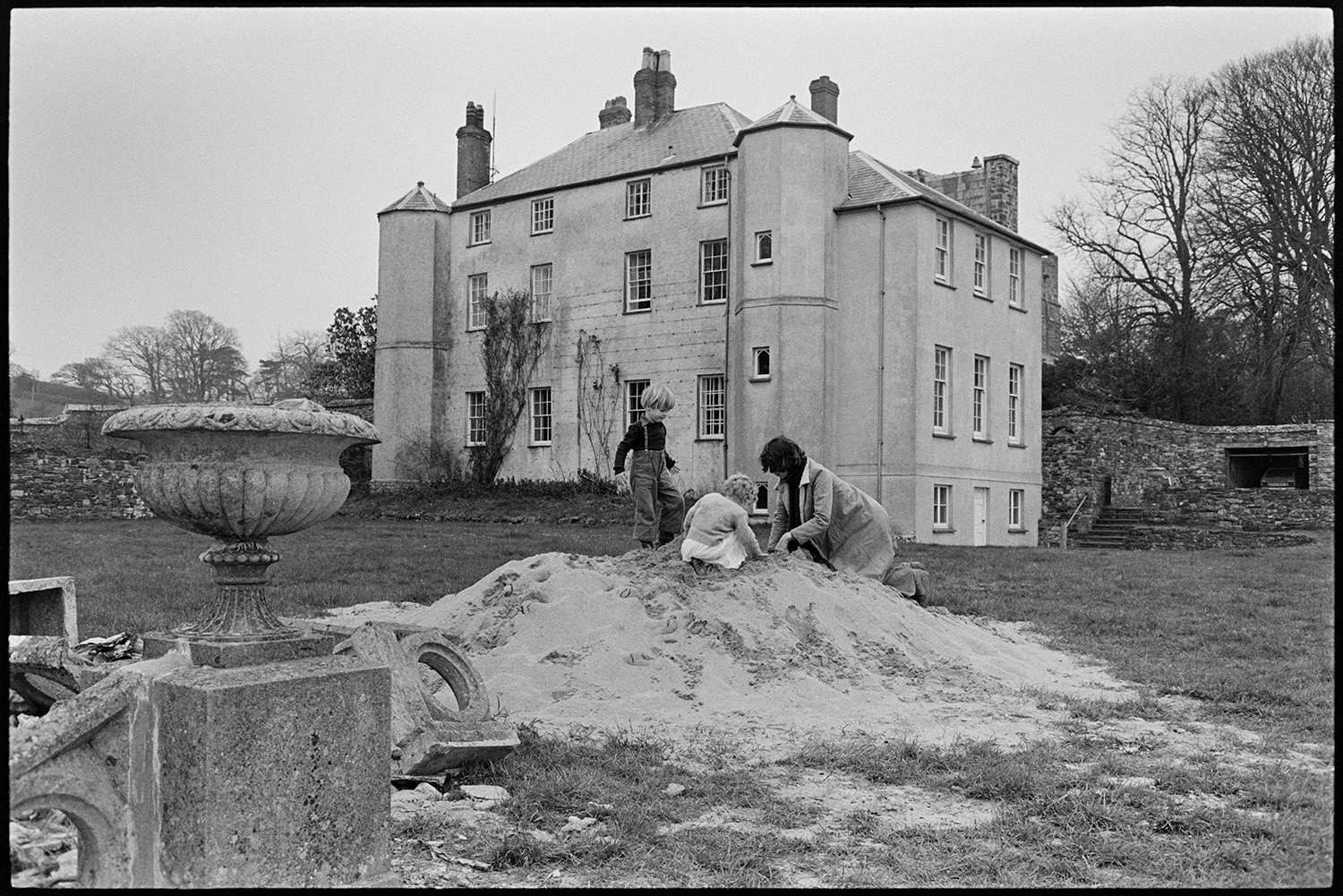 Christening, family tea and walk with children after service view of house, cliff walk.
[A woman and two young children from the Rous family playing in a pile of sand in the garden at Clovelly Court, after a Christening.]