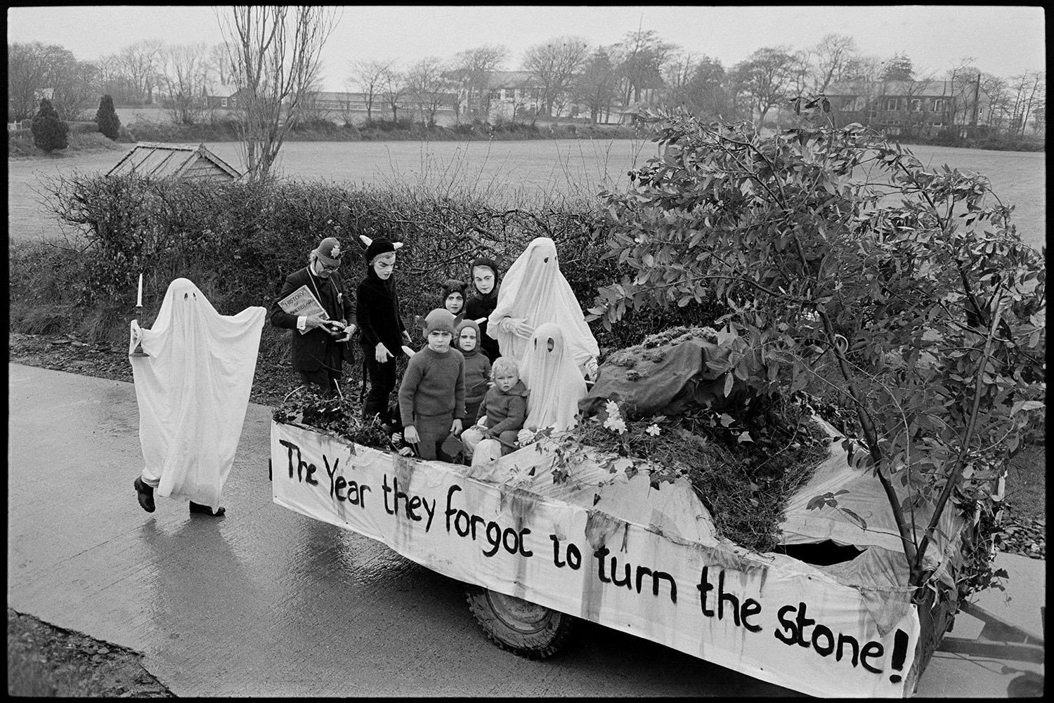 Carnival procession with Queen on decorated float, rain, spectators and Brass Band.
[Children dressed as ghosts and ghouls and a man dressed as a policeman on a carnival float in the Shebbear Carnival. The float is called 'The year they forgot to turn the stone!'.]