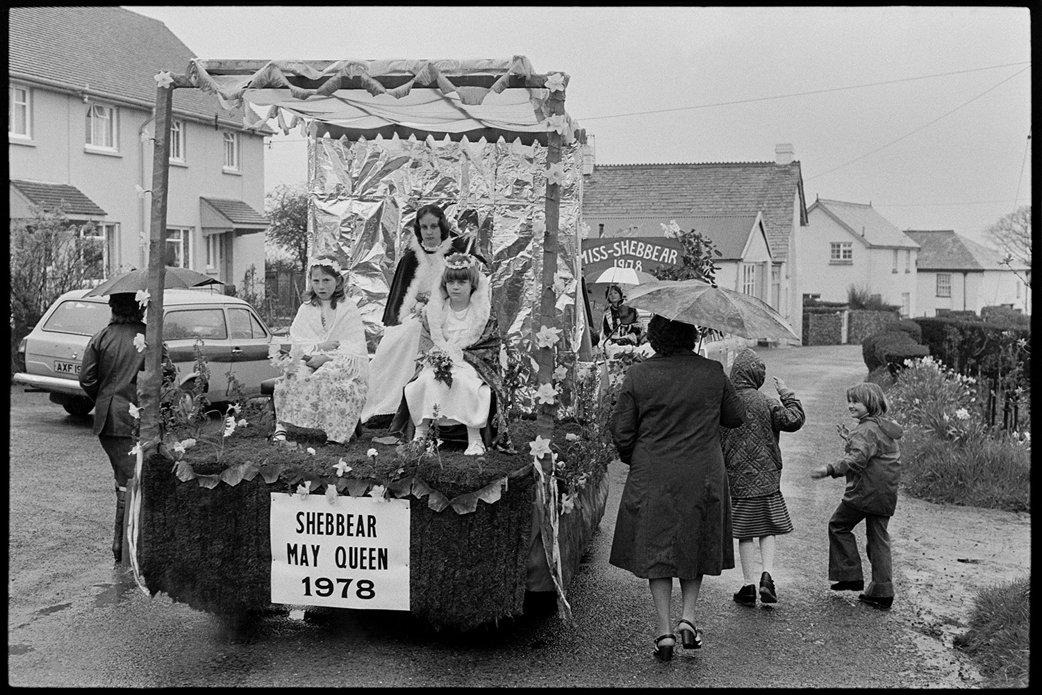 Carnival procession with Queen on decorated float, rain, spectators and Brass Band. 
[The Shebbear May Queen and two attendants sitting on a decorated trailer going to Shebbear Carnival. Miss-Shebbear is travelling on a trailer in front of the May Queen further along the street. Spectators are walking by the trailer holding umbrellas.]