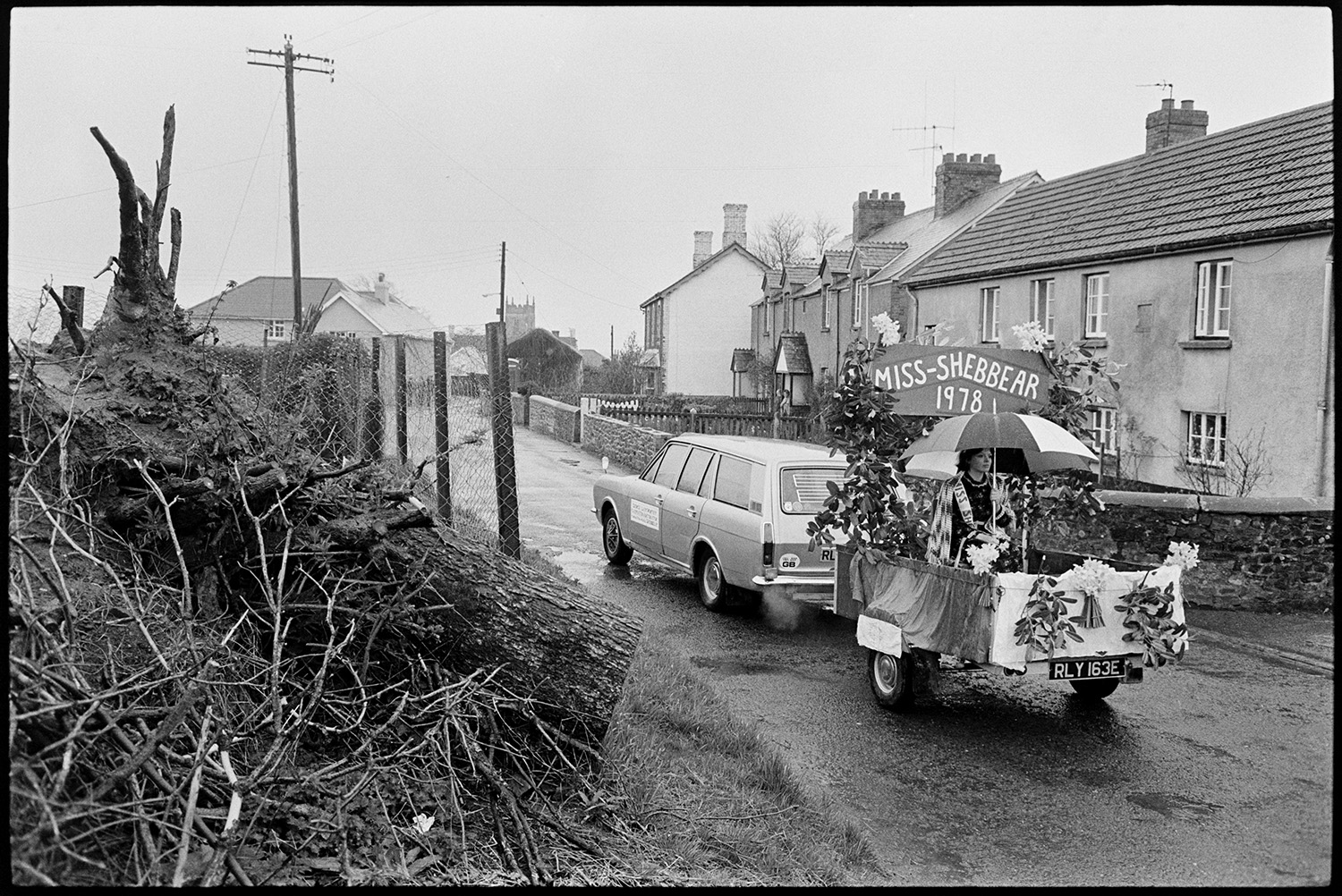 Carnival procession with Queen on decorated float, rain, spectators and Brass Band.
[A car pulling a trailer with Miss-Shebbear sitting under an umbrella driving along a road for Shebbear Carnival.]
