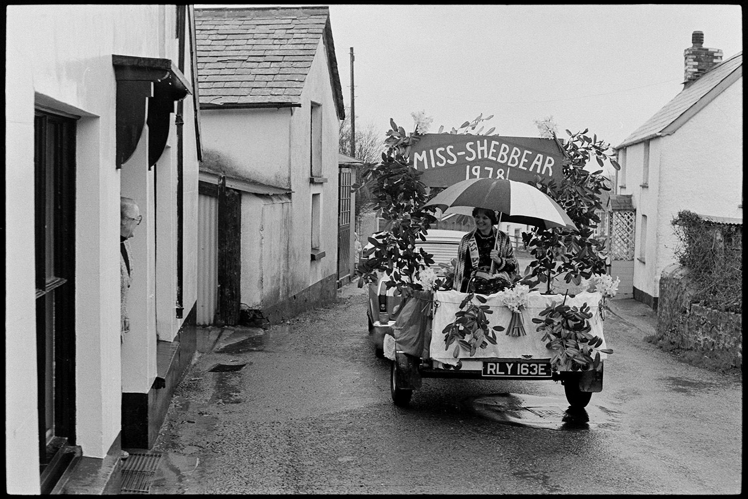 Carnival procession with Queen on decorated float, rain, spectators and Brass Band. 
[A car pulling a trailer with Miss-Shebbear sitting under an umbrella driving along a road for Shebbear Carnival. A person is watching her pass by from a front door of a house.]
