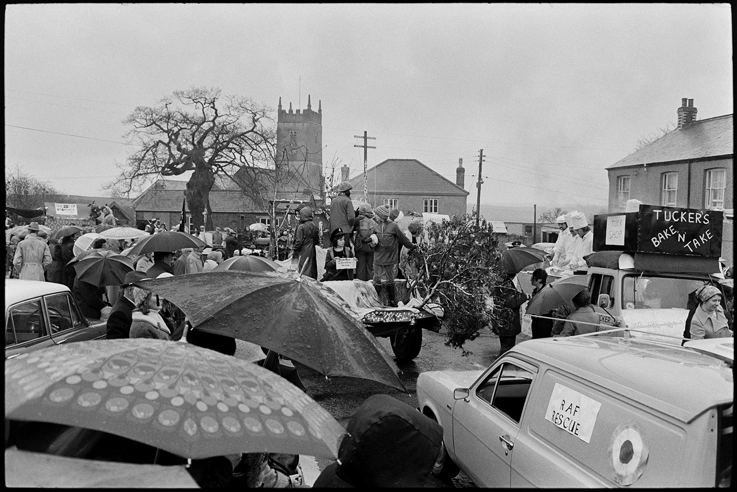 Carnival procession with Queen on decorated float, rain, spectators and fancy dress. 
[Spectators watching Shebbear Carnival in the rain, holding umbrellas. A carnival float is parading past and Shebbear church can be seen in the background.]