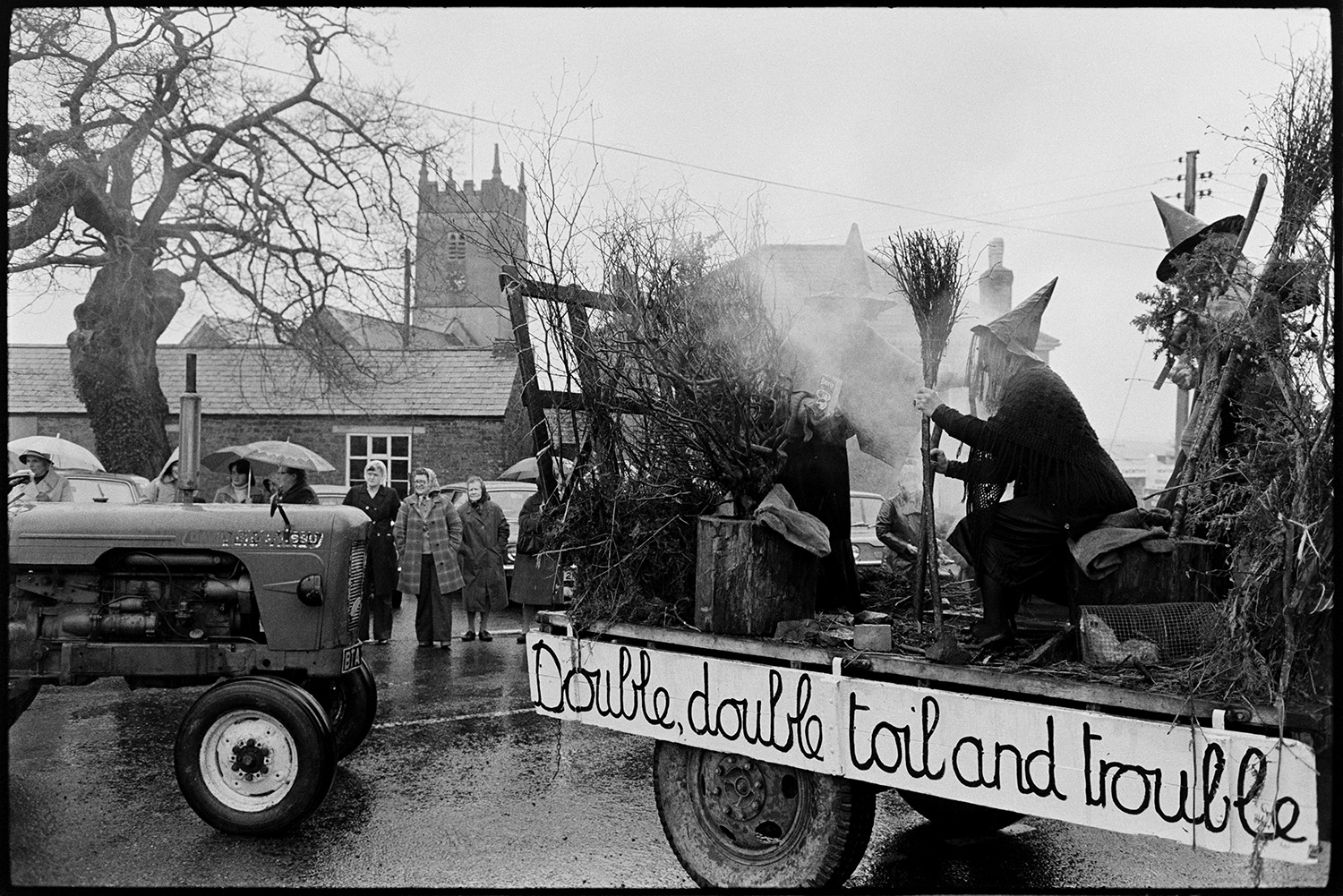 Carnival procession with Queen on decorated float, rain, spectators and fancy dress. 
[A carnival float in Shebbear Carnival with people dressed as witches. A banner on the side of the trailer reads 'Double, double, toil and trouble'. Spectators are watching the procession in the rain. Shebbear church tower can be seen in the background.]