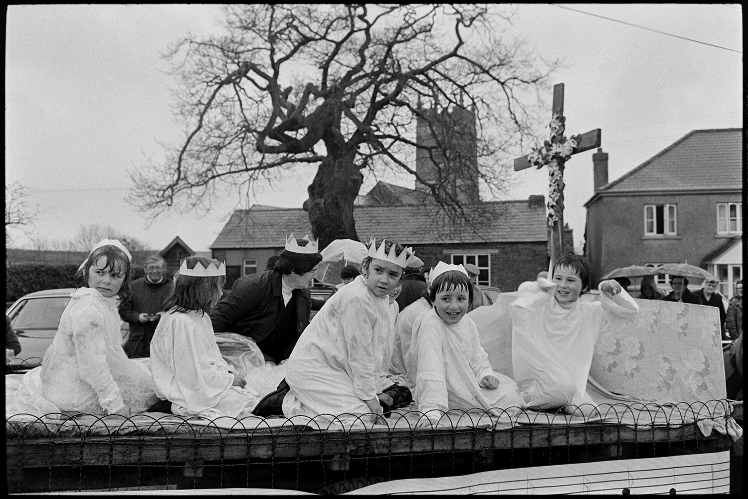 Carnival procession with Queen on decorated float, rain, spectators and fancy dress. 
[Children on a carnival float at Shebbear Carnival in fancy dress. Some of the children are wearing crowns and a cross with flowers is at the front of the float. Spectators are watching the procession in the rain with umbrellas. Shebbear church tower can be seen in the background.]
