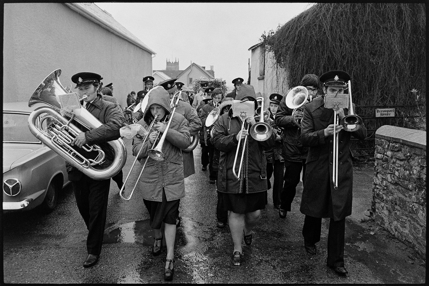 Brass band and fancy dress float at carnival marching through village. 
[A brass band marching along a street during Shebbear Carnival. The members are wearing coats to shield themselves from the rain.]
