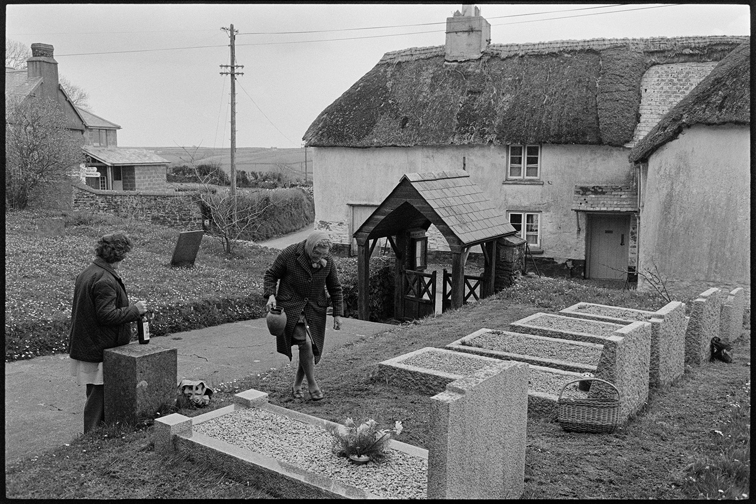 Two women tending graves with thatched farmhouse behind. 
[Two women in Roborough Churchyard. Mrs Wood is using a jug to water flowers on the graves. The lych gate entrance to the churchyard is visible. A thatched farmhouse can be seen in the background.]