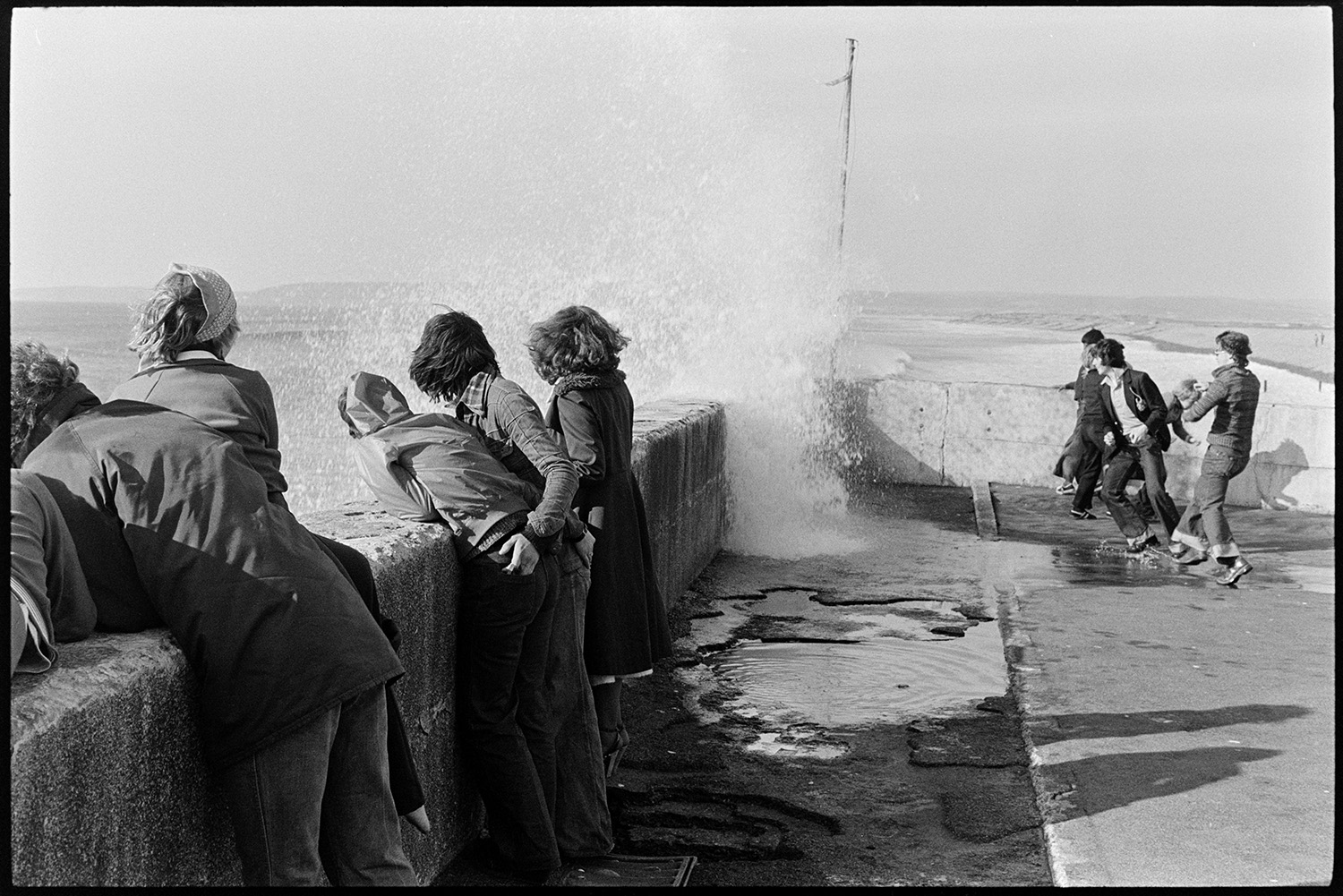 Young people dodging waves at sea front. 
[Teenagers dodging waves crashing over the sea wall at Westward Ho!]