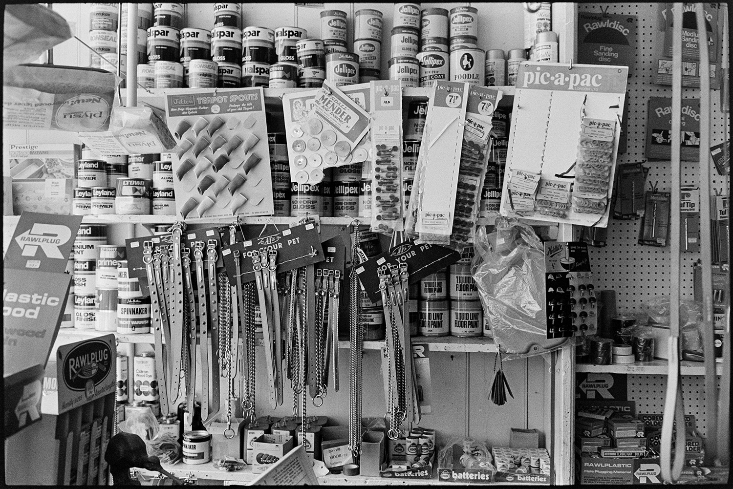 Shelves of stock, man serving paraffin with hand pump. 
[Items for sale in Eastmond's hardware shop in Torrington. Various goods are on display, including pet collars, teapot pours or spouts, tins of paint and batteries.]