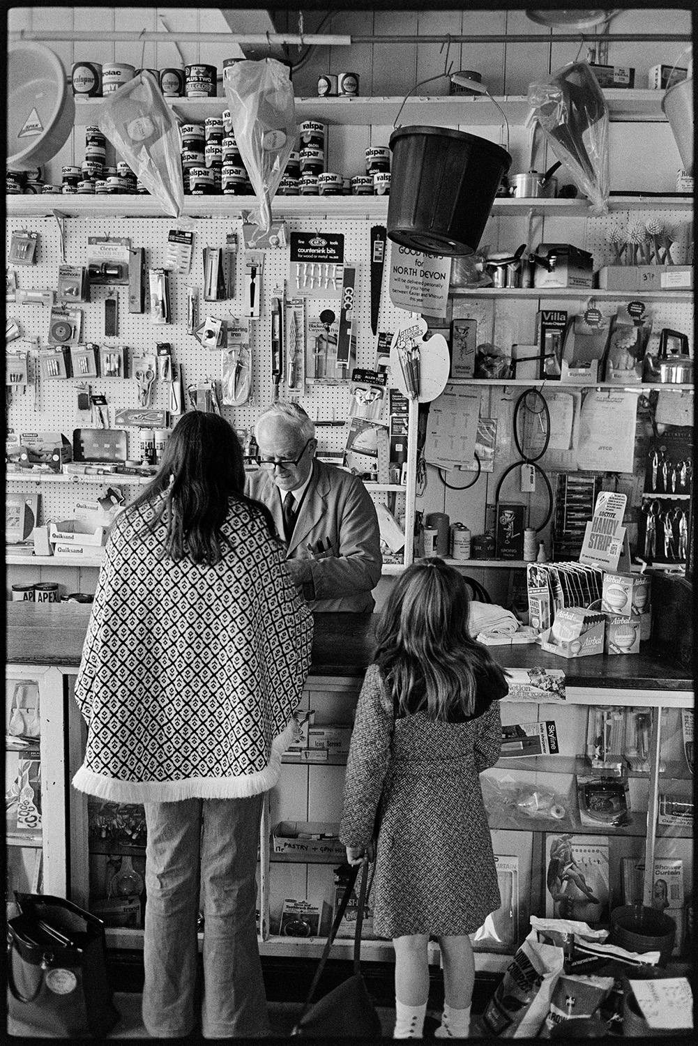 Shelves of stock, man serving paraffin with hand pump. 
[A man serving a woman and girl at the counter in Eastmond's hardware shop in Torrington. Various items are on display behind the counter including tins of paint, a bucket and scissors.]