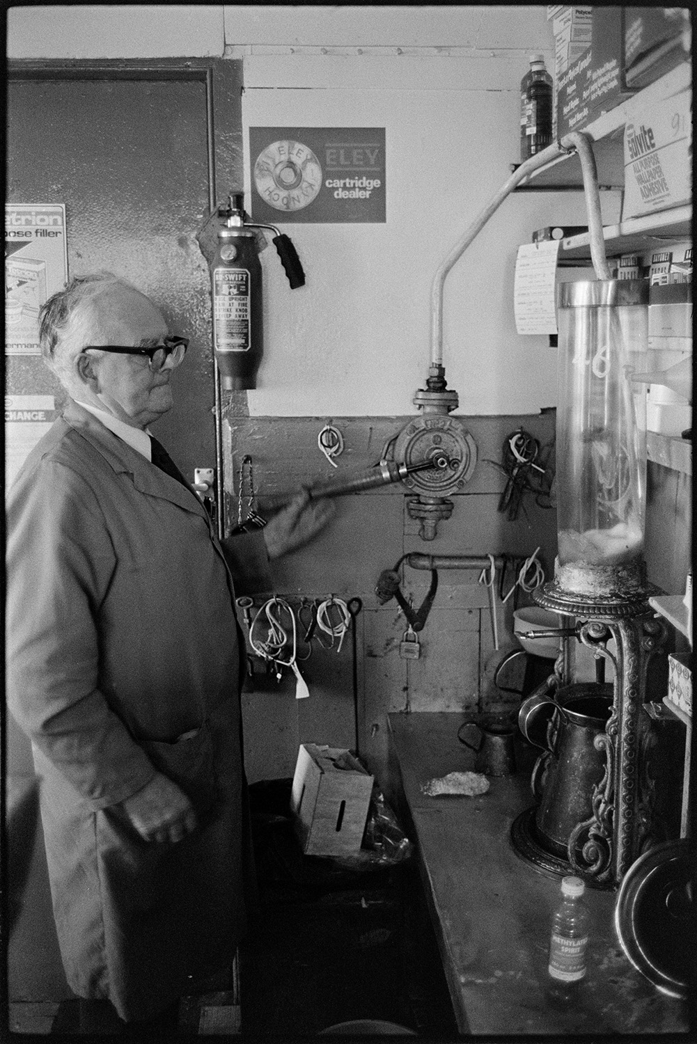 Shelves of stock, man serving paraffin with hand pump. 
[A man using a hand pump to measure out paraffin for a customer at Eastmond's hardware shop in Torrington.]