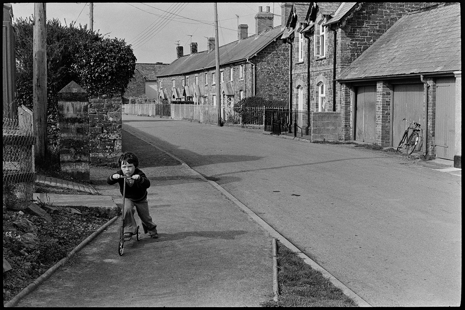 Street scene boy on scooter. 
[A boy pushing a scooter along a street in St Giles in the Wood. Garages and terraced houses can be seen in the background.]