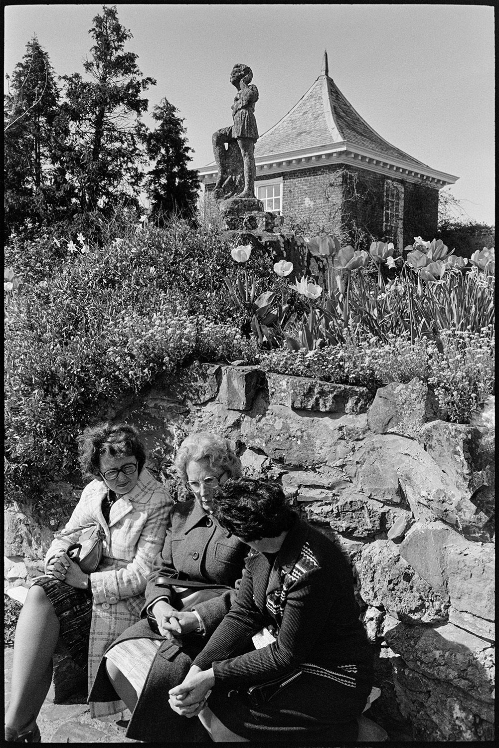 Garden party, open day visitors. 
[Three women sat on a bench talking at a garden party or open day in Fremington. A flower bed, statue and part of a building can be seen in the background.]