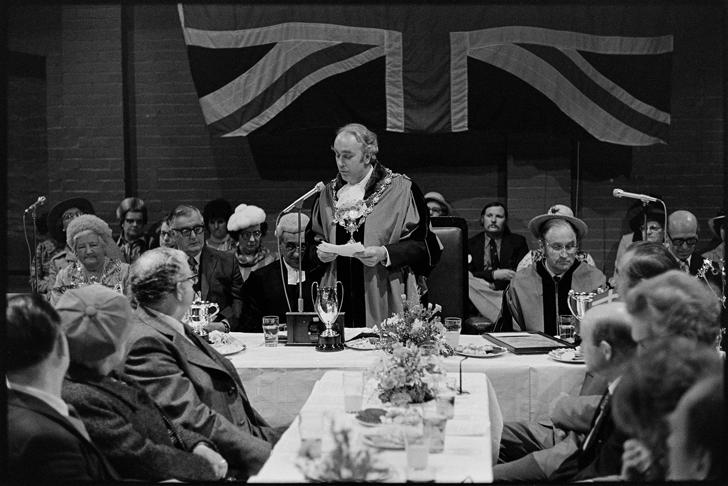 Mayors and dignitaries, Mayfair Banquet. Speeches, presentations and toasts to French twin town. 
[Dr Cramp, the mayor, making a speech at the Torrington Mayfair Banquet at The Plough, Torrington. Other dignitaries are sat behind him and a Union Jack flag is hung up in the background.]