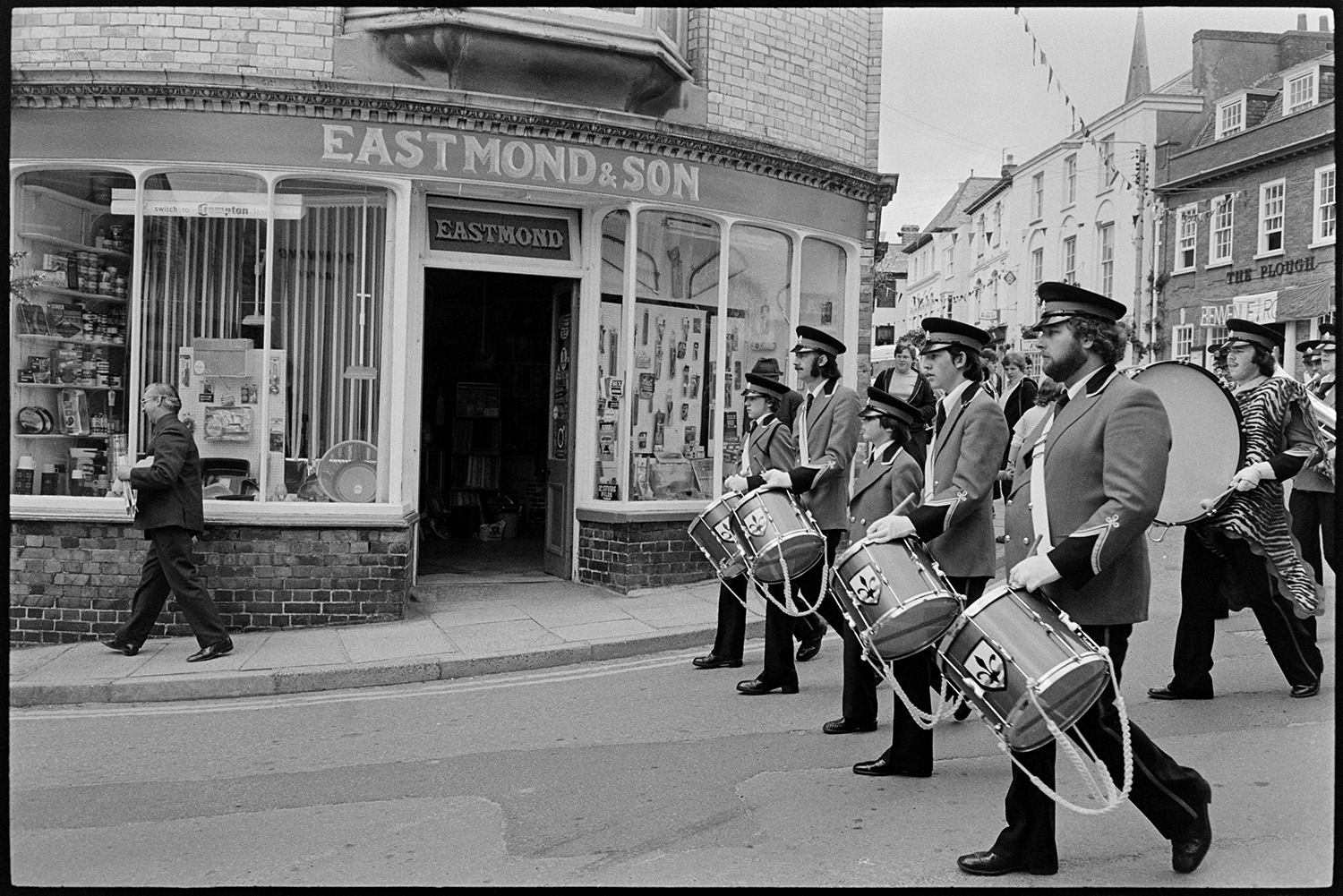 Mayors and dignitaries in parade round town making proclamation at points, town crier. 
[A marching band of drummers parading through Torrington for the Torrington Mayfair. They are passing the shop front of Eastmond & Son, hardware shop.]