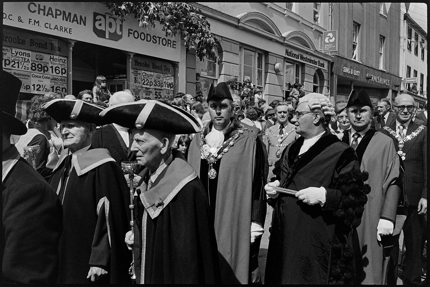 Mayors and dignitaries in parade round town making proclamation at points, town crier. 
[Dr Cramp, the Mayor, and other dignitaries parading through Torrington in the Torrington Mayfair parade. They are passing Chapman food store. Dr Cramp is wearing his mayoral chain of office and a bicorne hat. Spectators are lining the street watching the parade.]