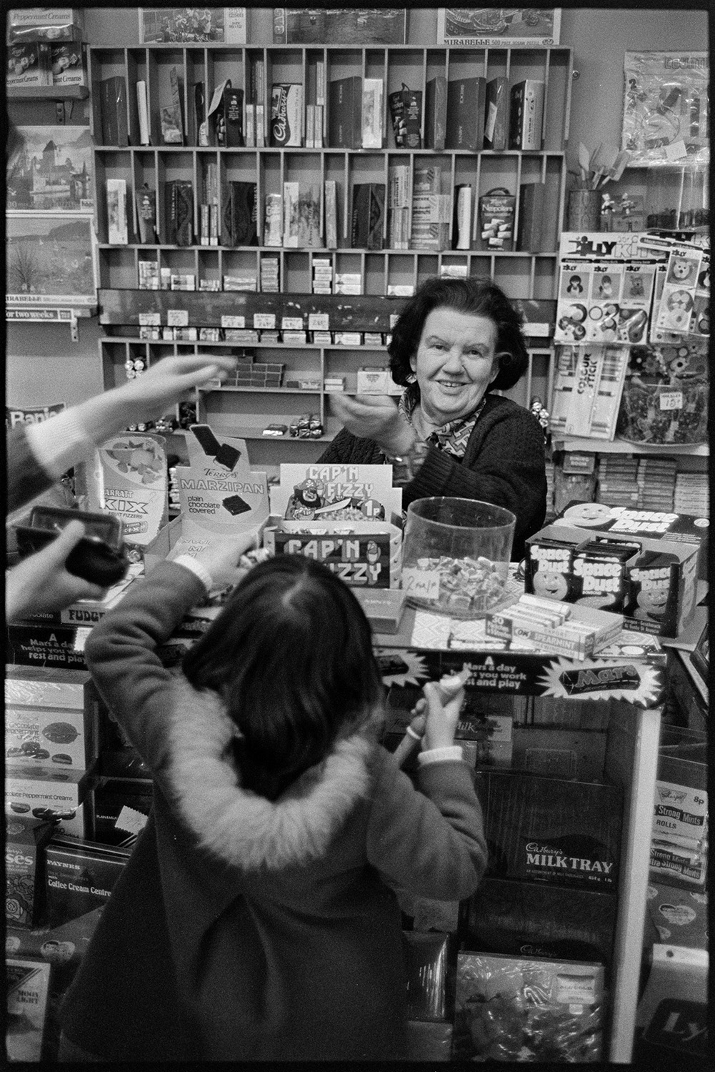 Woman serving children in her sweet shop. 
[A woman serving children in a sweet shop in The Square, Torrington. Sweets and chocolates are displayed on the counter and one of the children is holding a recorder.]