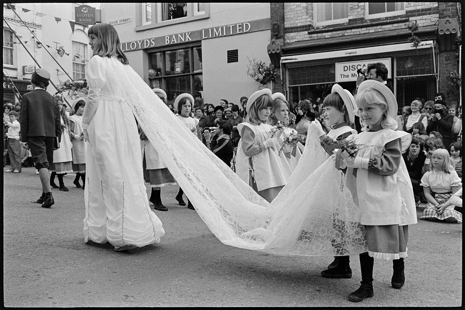 May Queen and Crowner processing to maypole, posing for photographers. 
[The May Queen and her attendants processing to the maypole for her to be crowned at Torrington Mayfair. Two of the attendants are holding the train of her dress. Spectators are watching from the side of the street outside Lloyds Bank.]