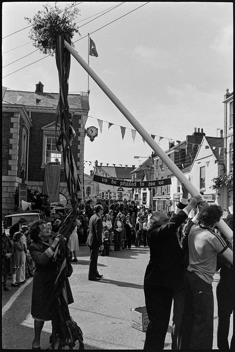 Hoisting the maypole in the town square. 
[Men hoisting the maypole in Torrington town square for Torrington Mayfair. A woman is holing the ribbons attached to the pole. Spectators are watching in the background and the street is decorated with bunting and banners.]