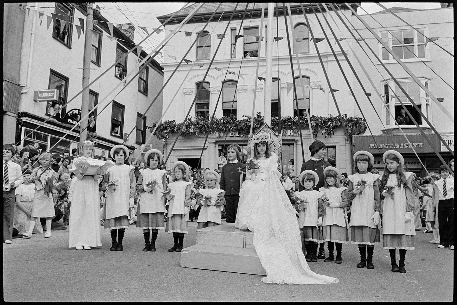 May Queen and Crowner processing to maypole, posing for photographers. 
[The May Queen and her attendants posing by the maypole at Torrington Mayfair for photographs. The May Queen has been crowned and is sitting on a throne. Tonya Copp is holding the cushion which the crown was presented on. Various buildings can be seen in the background, including an electrical shop and a pharmacy.]