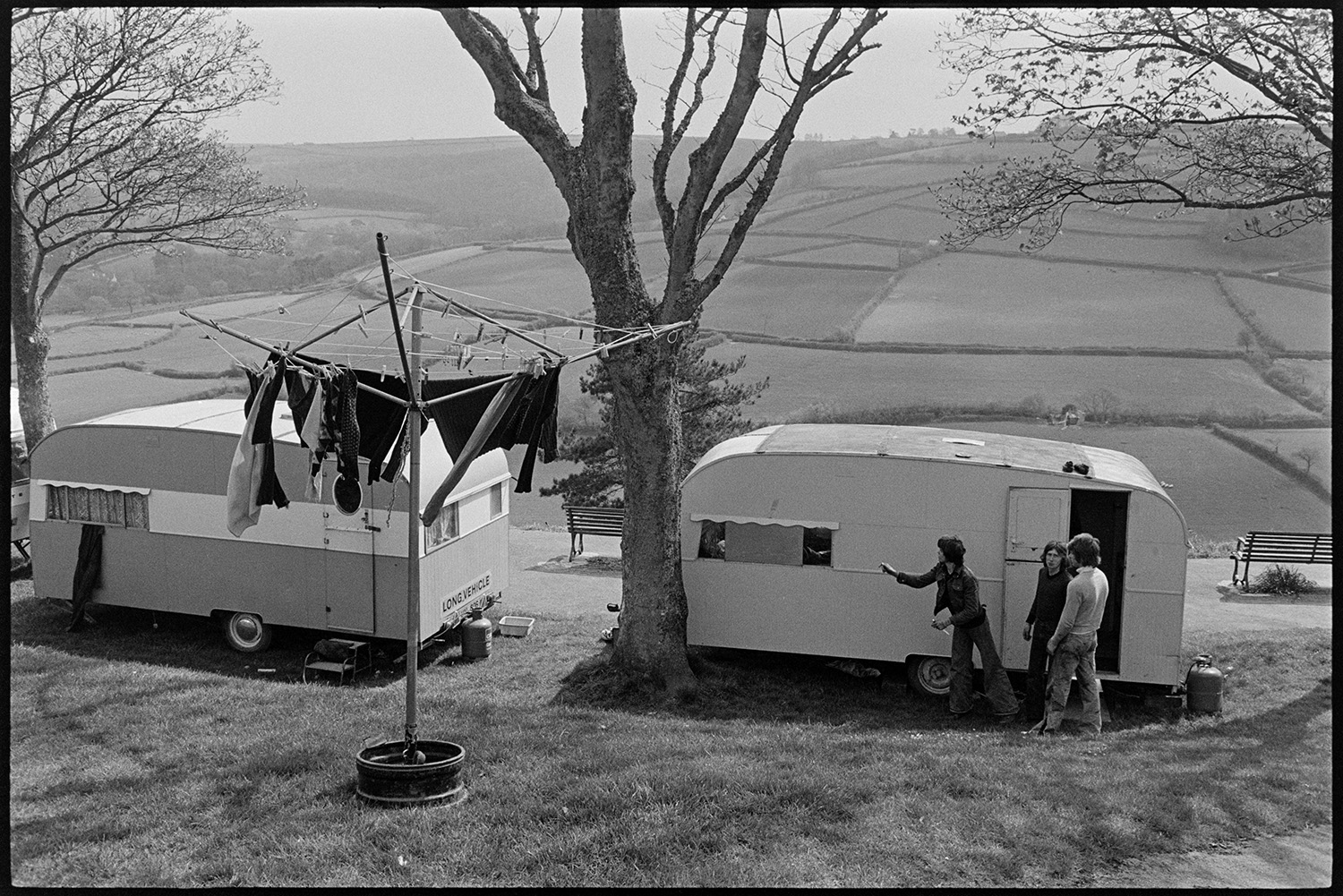 Two caravans with washing on clothes dryer, part of fair. 
[People outside two caravans which had come to Torrington Mayfair. They are parked in a park, possibly Castle Park, overlooking fields and hedges. Clothes are hung up to dry on a washing line in the foreground.]