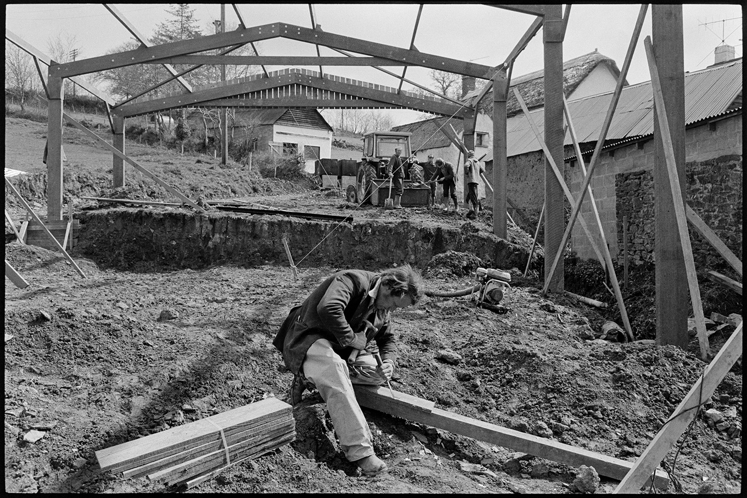 Men building metal and  timber framed barn shed. 
[Men building a barn with a timber and metal frame near New Bridge, Dolton. In the foreground a man is shaving a piece of wood with a chisel and hammer. In the background four men are stood by a tractor and link box. Other farm buildings are visible around the new barn.]