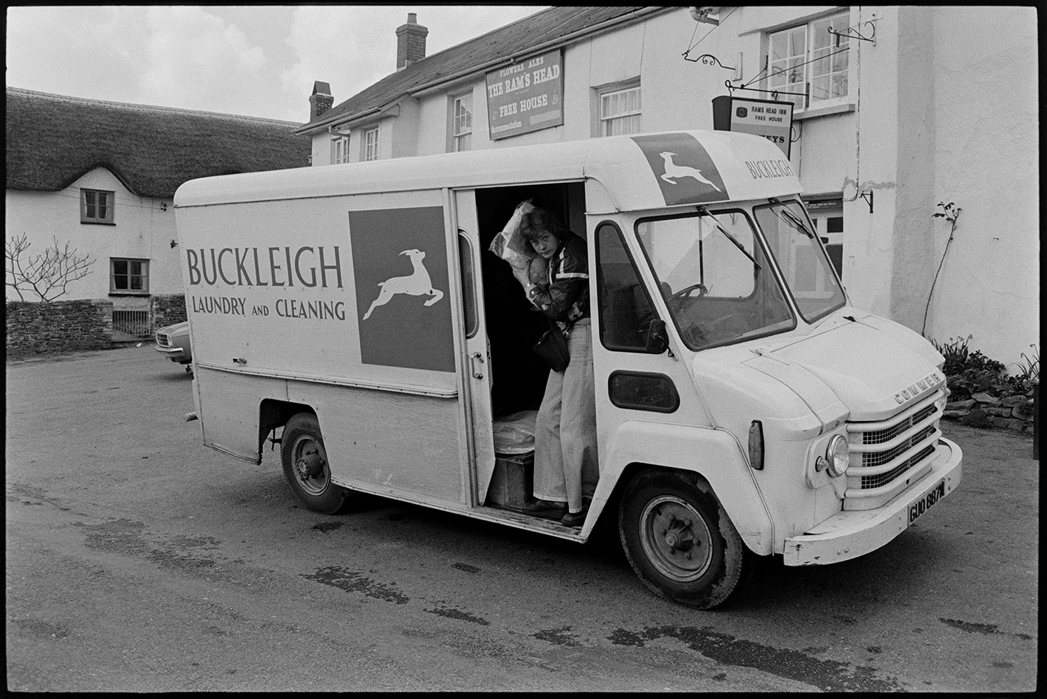 Laundry van parked in village, man delivering. 
[A person delivering laundry from a Buckleigh Laundry and Cleaning van, outside The Rams Head pub in Dolton.]