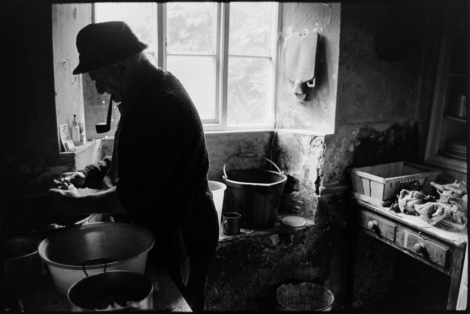 Farmer peeling potatoes and preparing meal in kitchen. Saucepans, vegetables, etc. 
[Archie Parkhouse peeling potatoes and placing them in a saucepan for a meal in his kitchen at Millhams, Dolton. He is smoking a pipe. A hot water bottle is hung up on the wall.]
