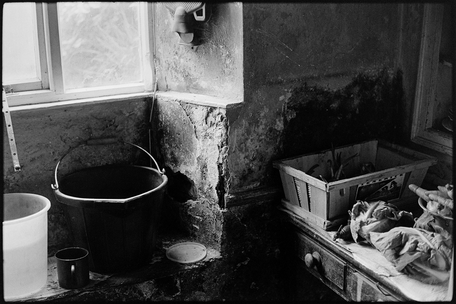 Farmer peeling potatoes and preparing meal in kitchen. Saucepans, vegetables, etc. 
[Archie Parkhouse's kitchen at Millhams, Dolton. Vegetables are on a wooden table with drawers, a bucket is placed in the window sill and a hot water bottle is hung on the wall.]