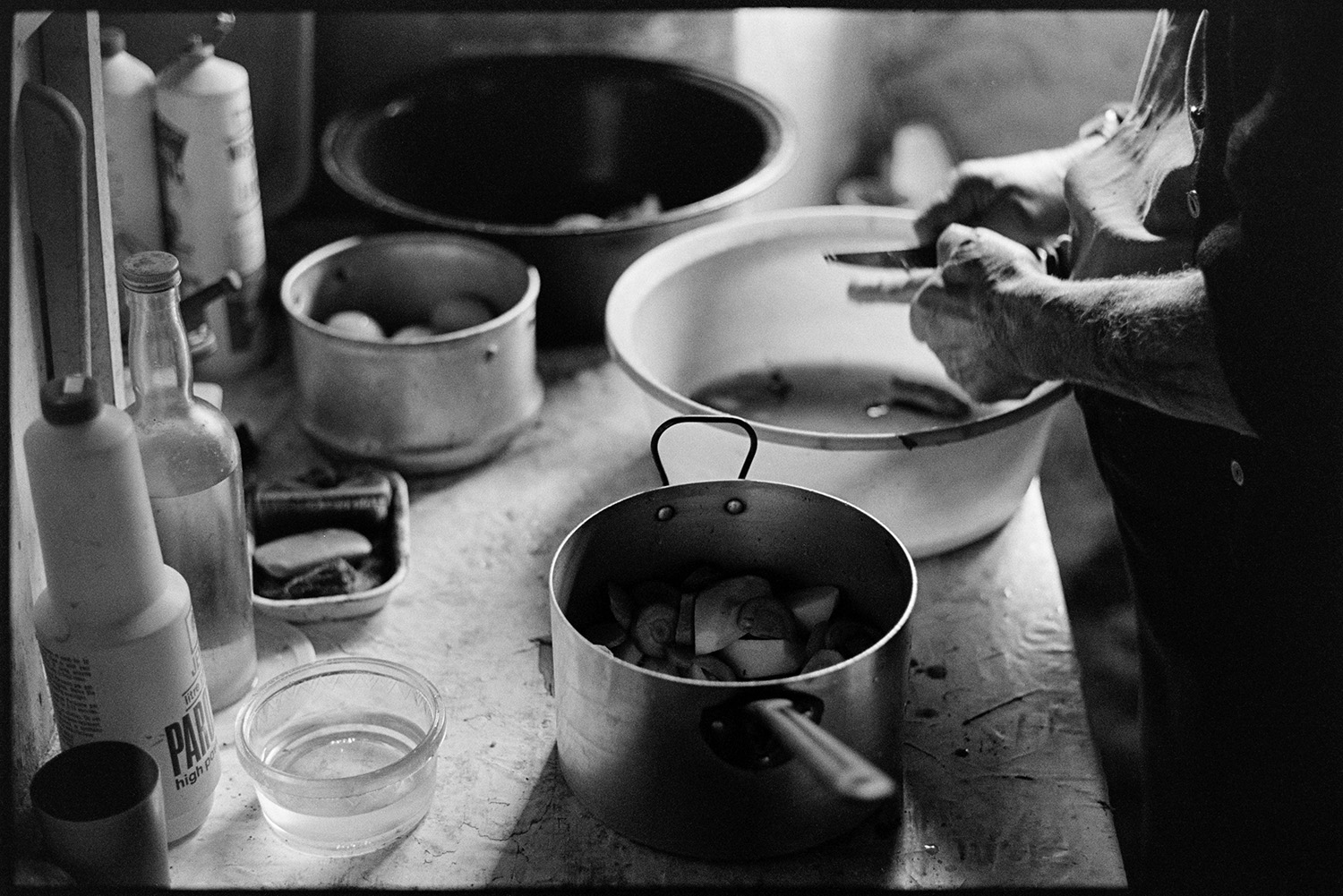 Farmer peeling potatoes and preparing meal in kitchen. Saucepans, vegetables, etc. 
[Archie Parkhouse peeling vegetables and placing them into a saucepan in his kitchen at Millhams, Dolton. Other pots and pans are visible, as well as various bottles and a bar of soap.]