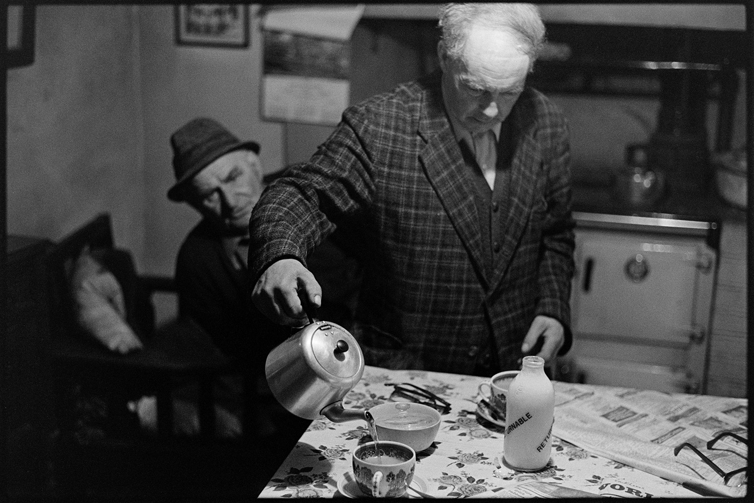 Man pouring tea. 
[Ivor Brock pouring tea from a teapot into a cup on his kitchen table at his cottage at Millhams, Dolton. A bottle of milk is also on the table. Archie Parkhouse is sat in the background by a rayburn stove.]