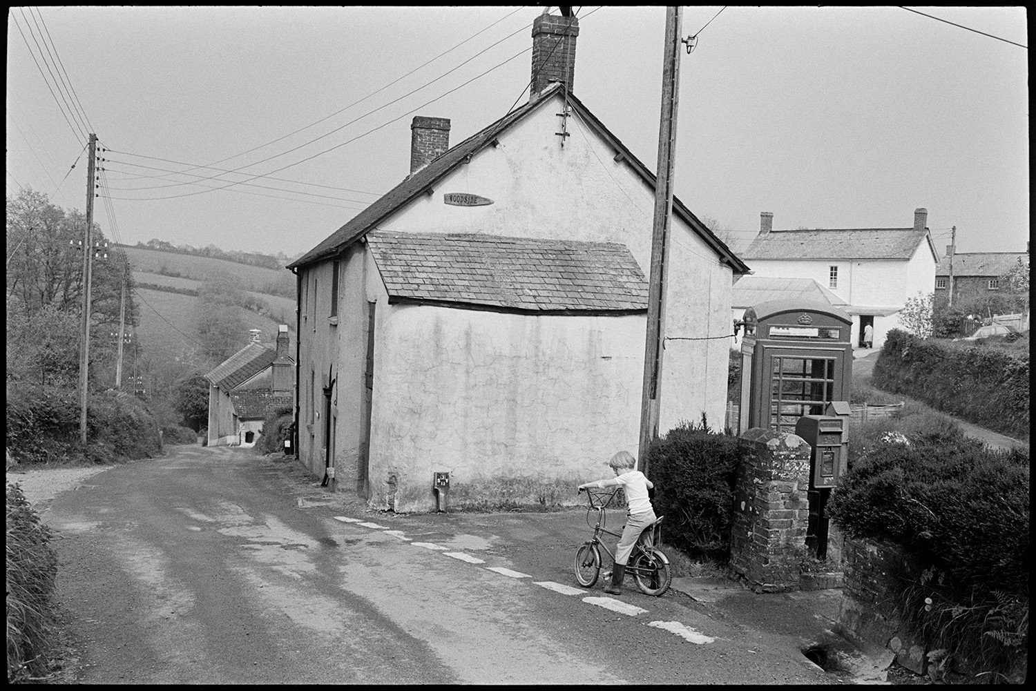 Street scene, child on bicycle. 
[A child riding a bicycle past a telephone box, post box and houses in Hollocombe.]