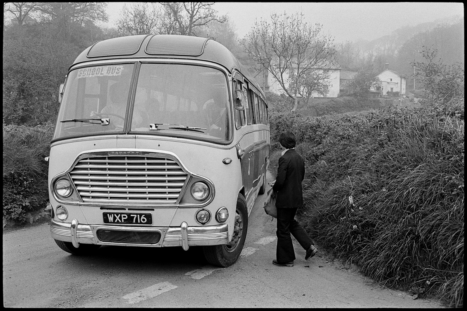 Boy waiting for school bus and getting into it. 
[A boy getting onto the school bus in Hollocombe. A house is visible in the background.]