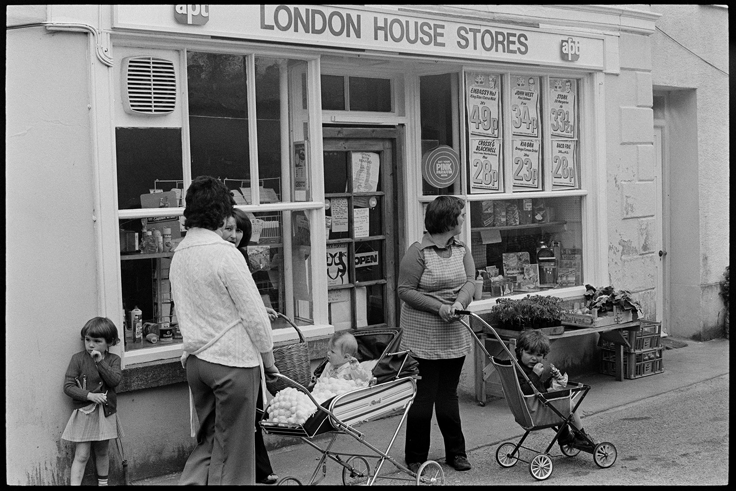 Mothers, women and children with prams outside shop. 
[Women with prams and children talking outside the shop front of London House Stores in Fore Street, Dolton. Various adverts can be seen in the shop window and vegetables are displayed outside.]