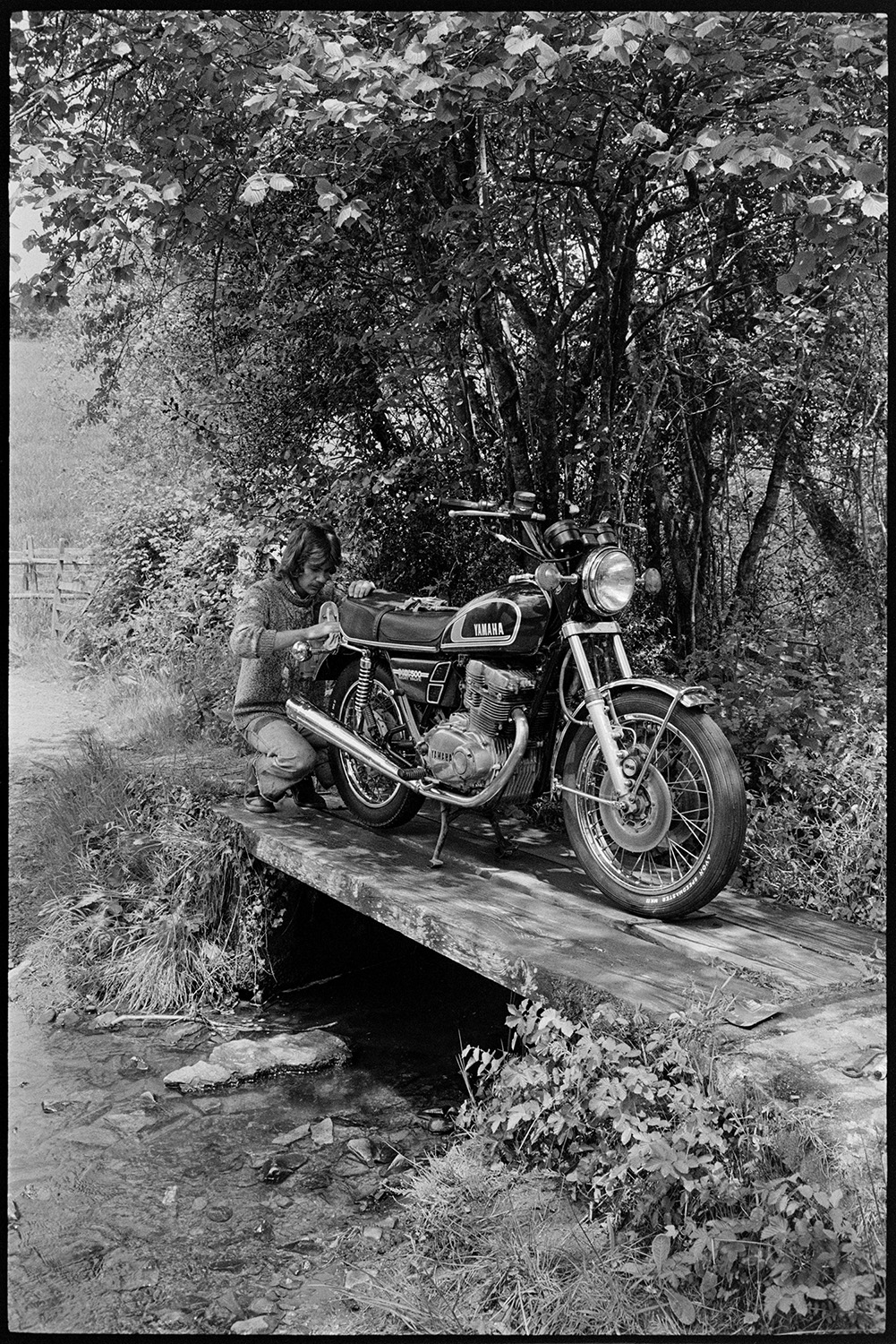 Man cleaning motorbike prior to sale. 
[A man cleaning his motorcycle on a small wooden bridge over a stream at Addisford, Dolton, before selling it.]