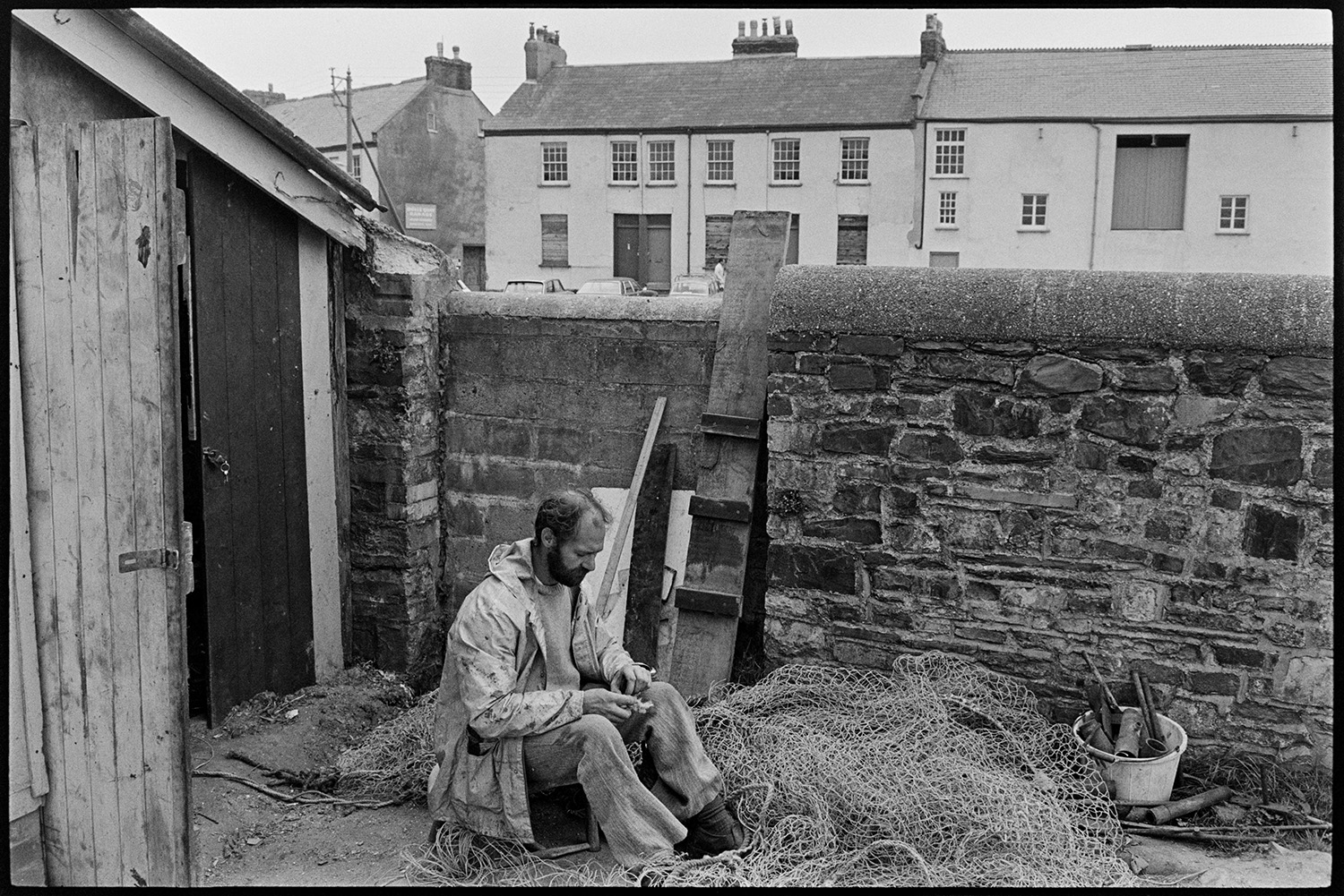 Fisherman mending nets on quay. 
[A fisherman mending his nets by a shed and stone wall on Rolle Quay, Barnstaple.]