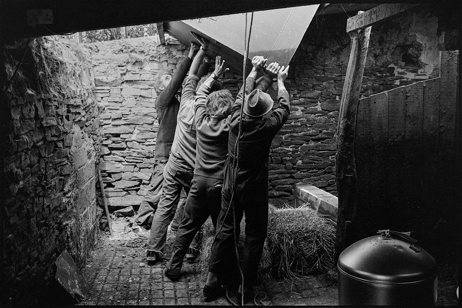 Men heaving large oil tank into shed. 
[Four men lowering a large oil tank into a stone barn at Woolridge, Dolton. They are lowering it onto hay bales.]