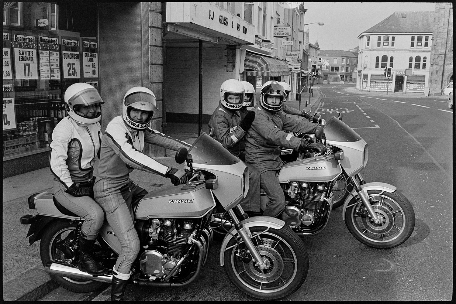 Motorcyclists parked in town. 
[People on motorbikes parked on the side of a road in Okehampton. Shop fronts can be seen in the background.]
