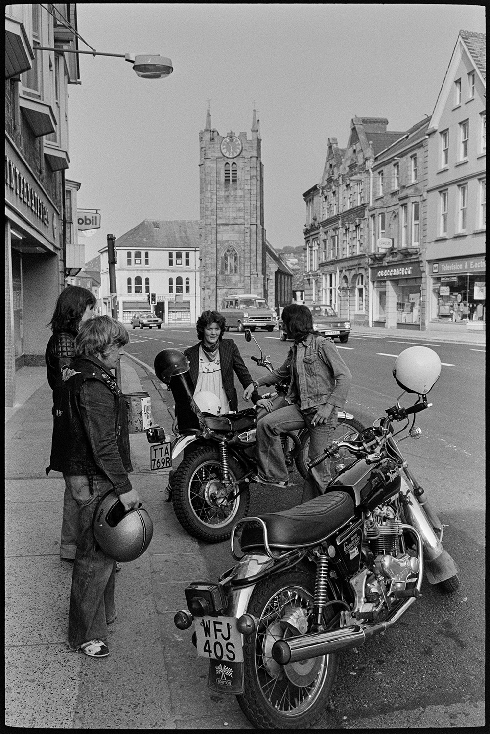 Motorcyclists parked in town. 
[Motorcyclists talking by their motorbikes which are parked on the side of a road in Okehampton. Shop fronts and a church tower can be seen in the background.]