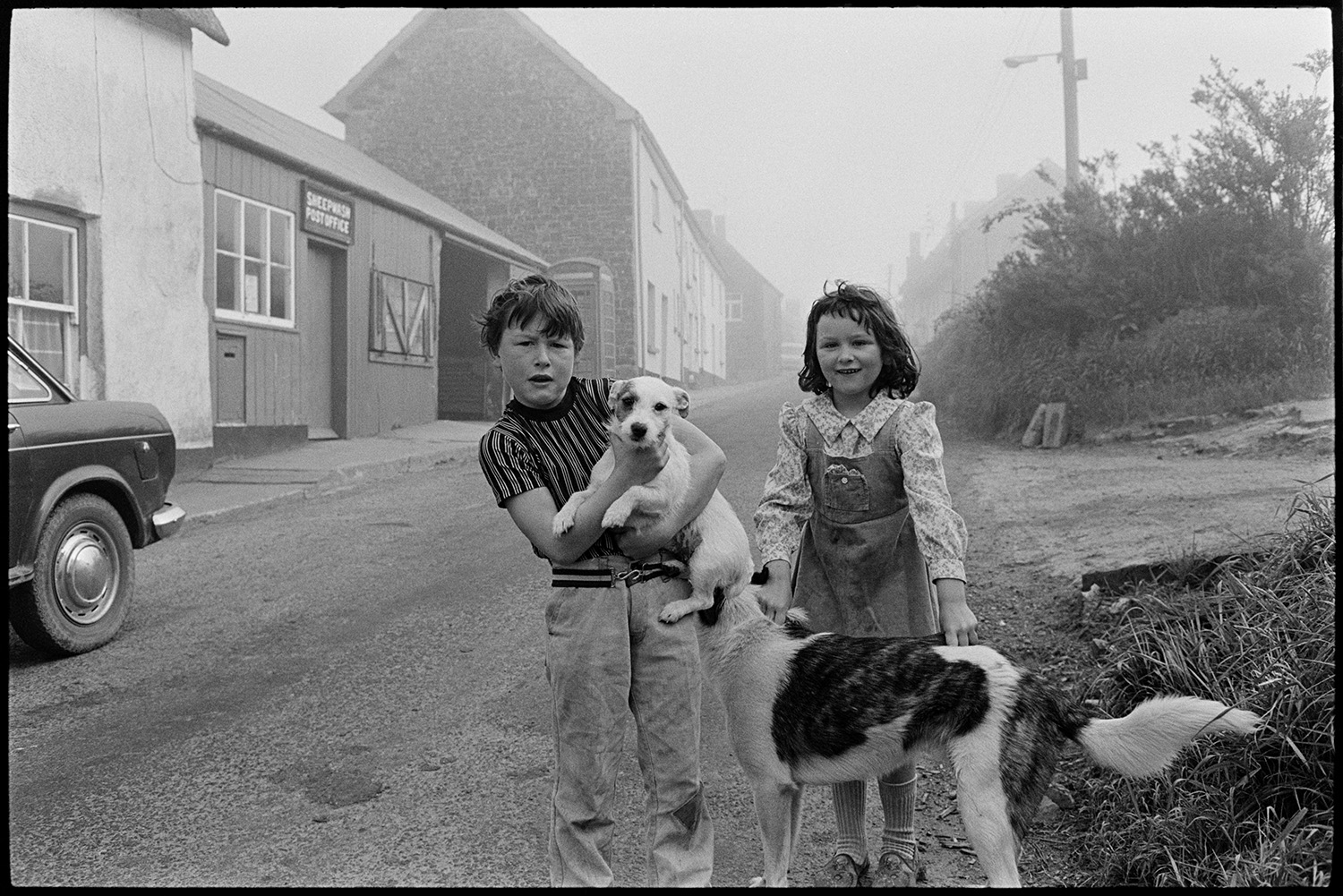 Street scenes with children, car and telephone kiosk. 
[A boy and girl, and two dogs, playing in the street outside Sheepwash Post Office, in Sheepwash. A telephone box and car can be seen in the background.]