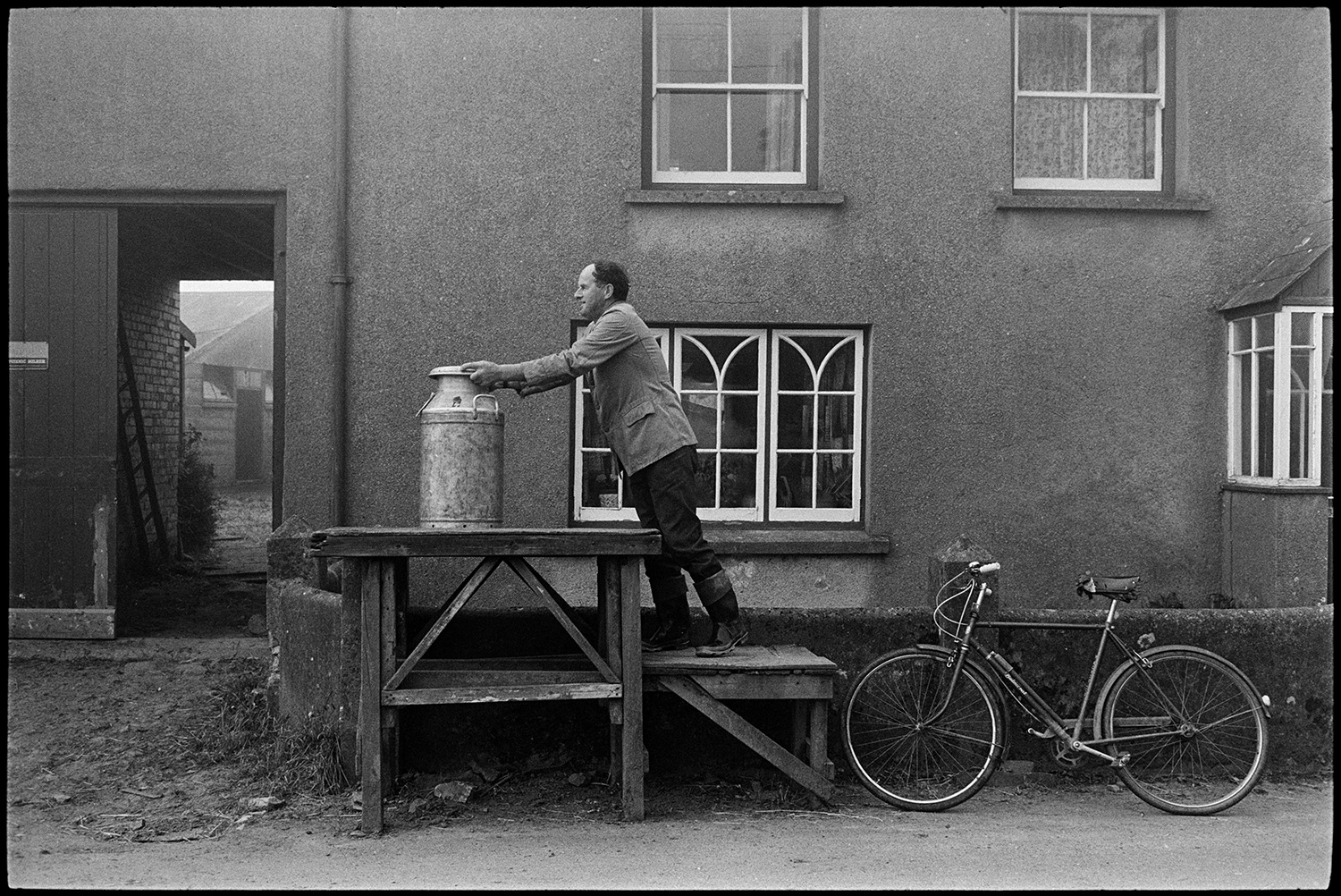 Farmer rolling out milk churn and putting it on stand. 
[Walter Newcombe placing a milk churn on a wooden milk churn stand outside a house with decorative windows, possibly his farmhouse, in East Street, Sheepwash. His bicycle is resting against a wall outside the house.]
