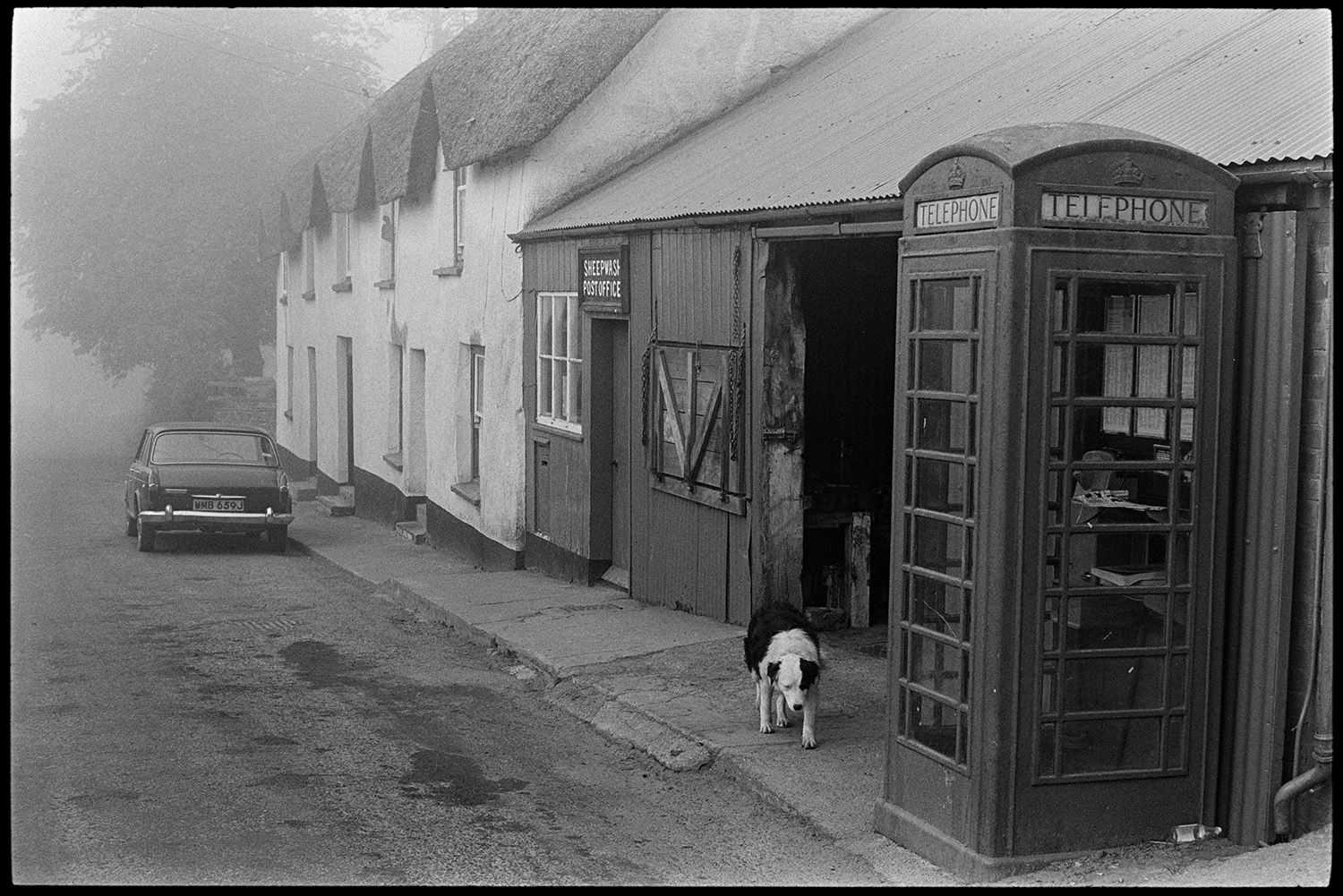 Street scenes with children, car and telephone kiosk. 
[A dog walking past a telephone box and Sheepwash Post Office, at Sheepwash. Thatched cottages and a car are visible further along the street.]