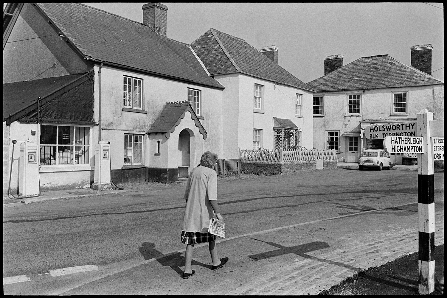 Village shop inside and out. 
[A woman walking past the village shop at Sheepwash. Two petrol pumps are outside. A signpost is in the foreground and houses can be seen in the background.]