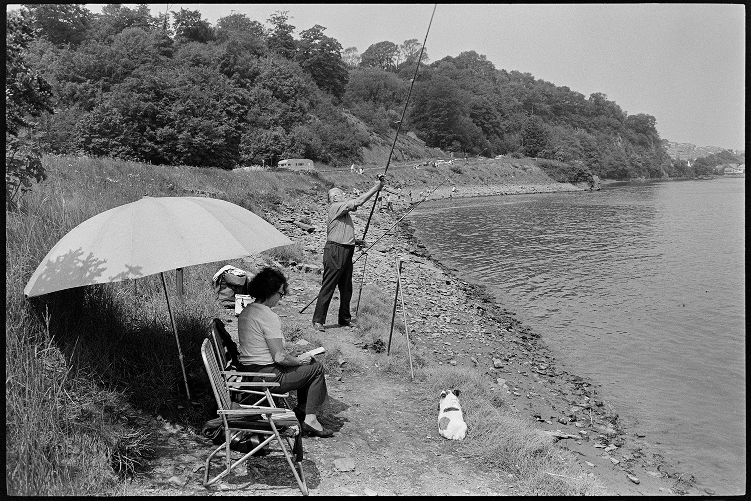 People fishing and having picnic beside estuary, tourists. 
[A man fishing on the River Torridge estuary at Bideford. A woman is sat reading a book under a large parasol, and a dog is sitting at the estuary edge watching the man fishing.]
