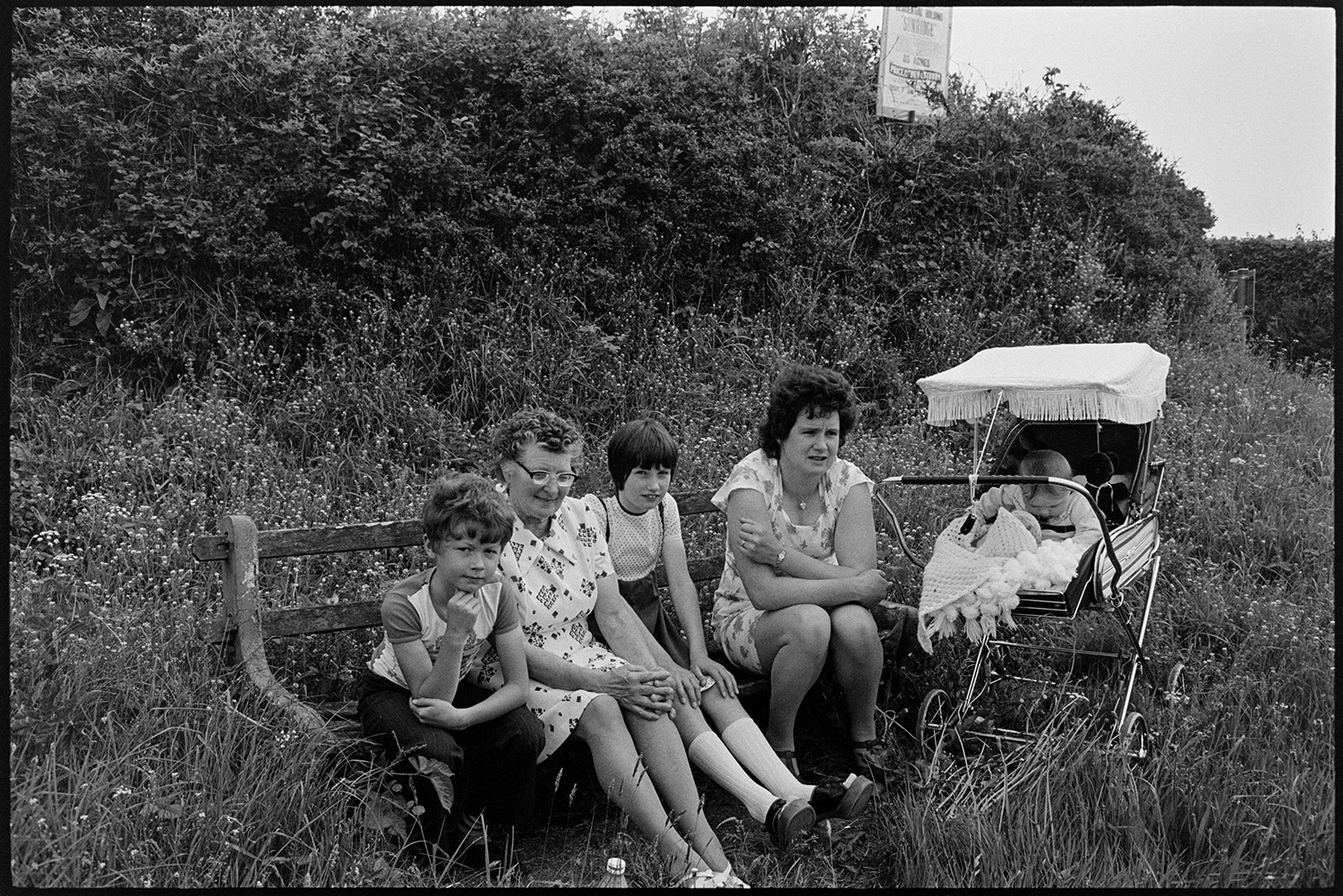 Family sitting beside road junction, now altered. 
[Two women and two children sat on a wooden bench by a roadside at Beaford. A pram with a baby is also with them. The bench is surrounded by foliage and a hedge.]